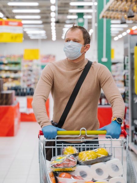 Man walks in supermarket with shopping trolley with goods, wears medical mask